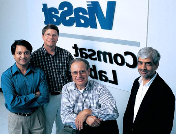 Four Viasat employees smiling at the camera in front of a Viasat Comsat Lab sign