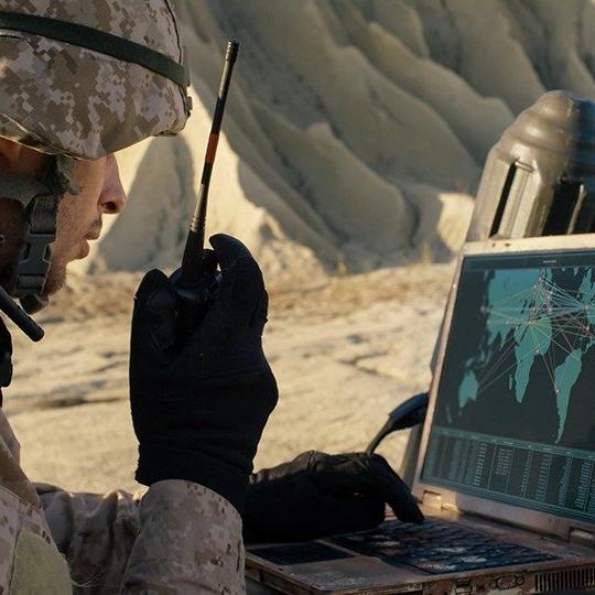 一名身穿战斗装备的男子一边对着收音机讲话，一边看着军用笔记本电脑上的世界地图